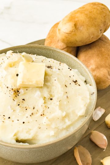 A tan bowl full of dairy free creamy garlic mashed potatoes. A pat of butter, salt and pepper as garnish. In the background garlic cloves and unpeeled potatoes.