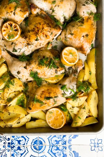 Cooked Chicken and potato wedges coated in herbs in a large sheet pan on a blue and white tiled counter.