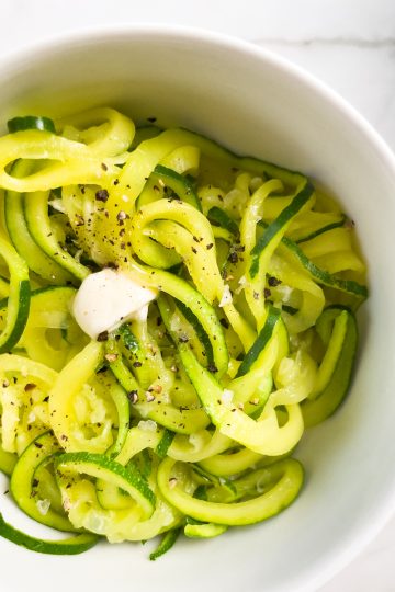 Zucchini Noodles in a white bowl with salt and pepper and a pat of dairy free butter melting on them.