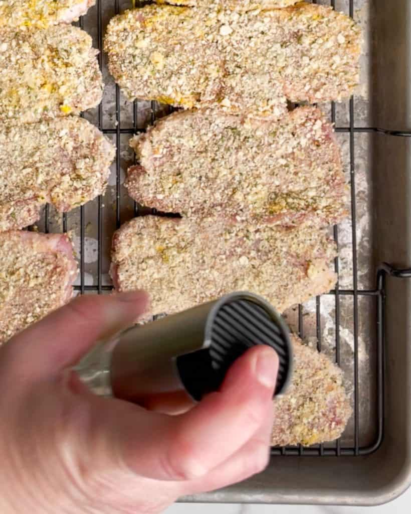 gluten free baked pork schnitzels on a rack with a hand holding an oil sprayer spraying them with oil.
