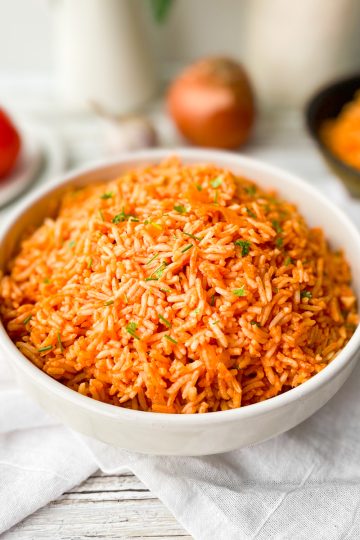 Mexican vegan rice in a white bowl with tomatoes on a plate to the left behind and a skillet with fried rice behind and to the left.