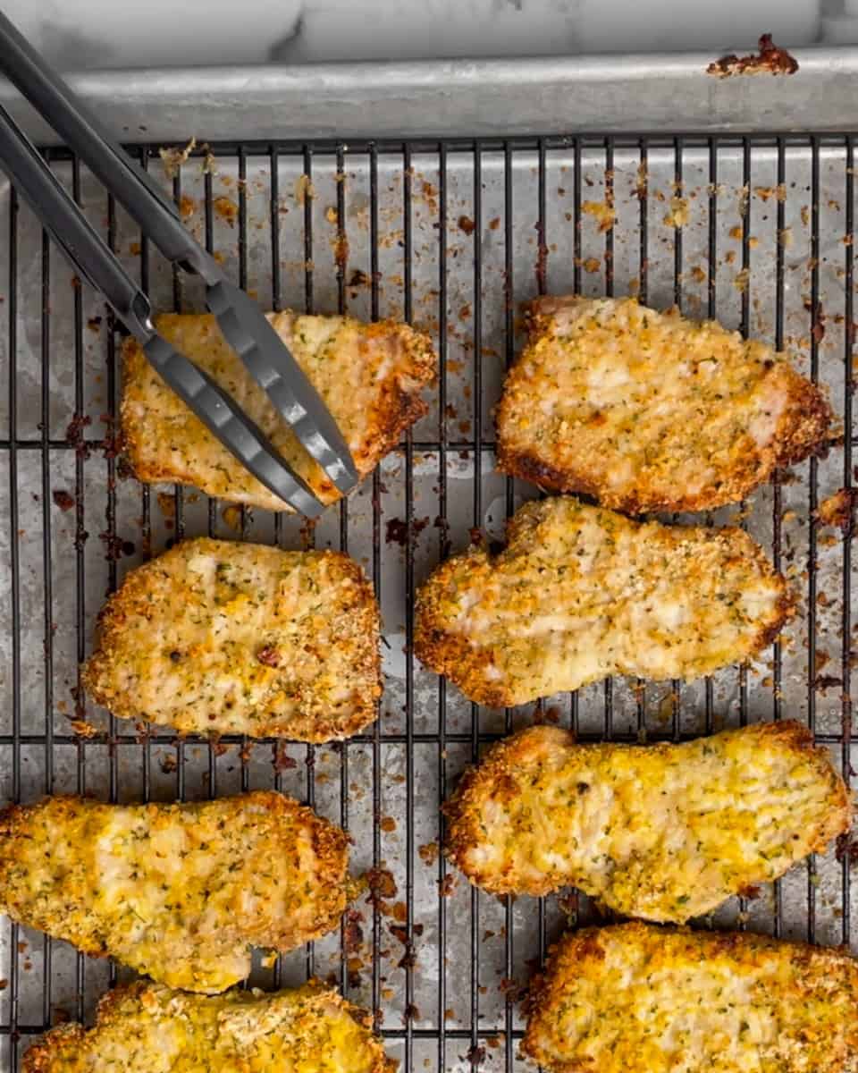 a pair of tongs resting on a pork chop in the top left corner and baked pork schnitzels on a rack.