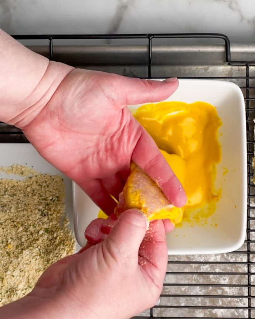 Two hands showing how to remove the excess mustard format he pork chop. One hand gripping the end of the pork chop and pulling away from the mustard bowl and the other hand placing a finger on either side lightly to remove the mustard.