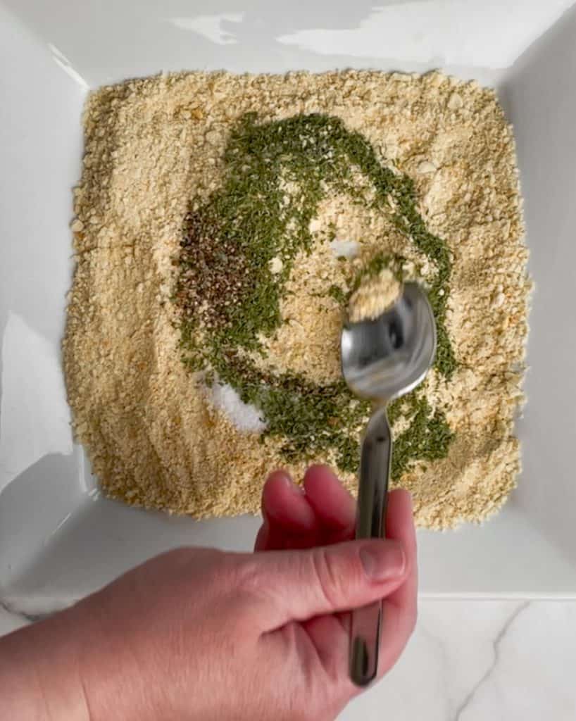 A hand holding a spoon and mixing the herbs and spices into the bread crumbs.