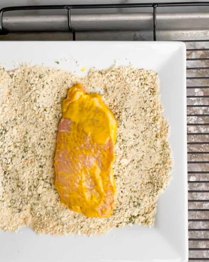 A pork chop coated in mustard laying on a bed of bread crumbs on a shite plate.