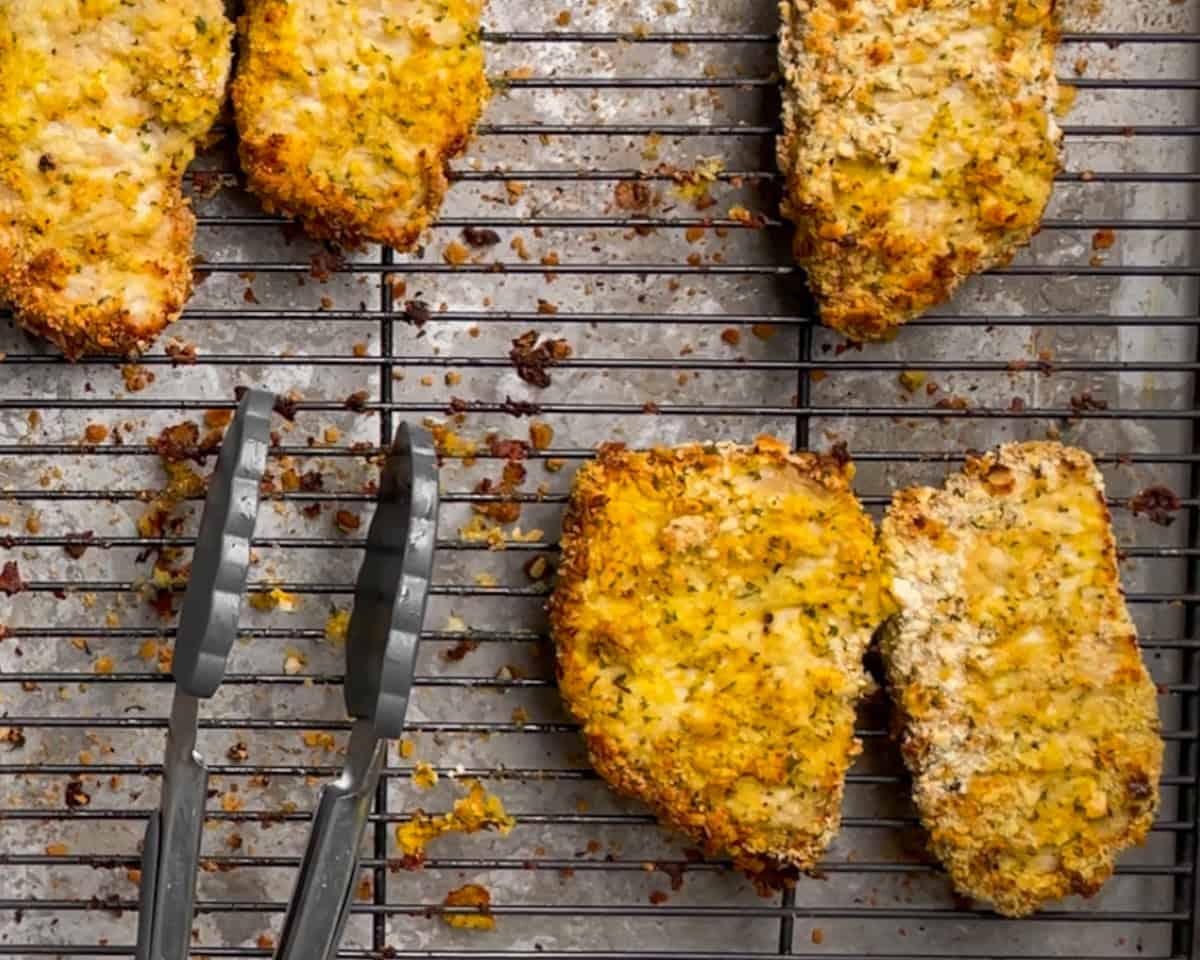 a pair of tongs resting in the bottom left corner and baked pork schnitzels on a rack.