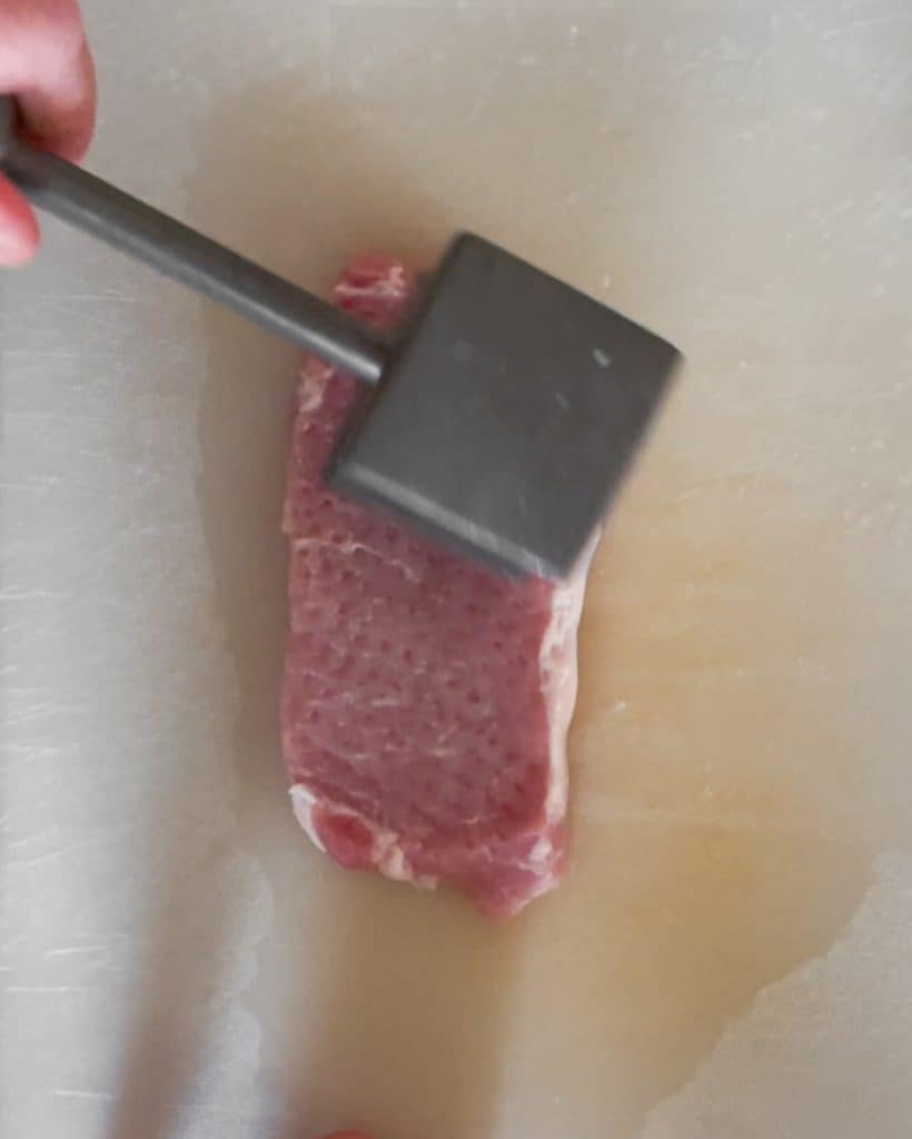 A hand holding a meat mallet with the flat side up. The textured side of the mallet is being used on the pork chop on a white plastic cutting board.