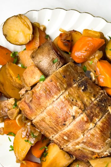 A white platter with a whole pot roast in the centre surrounded by cooked potatoes, carrots, and onions.